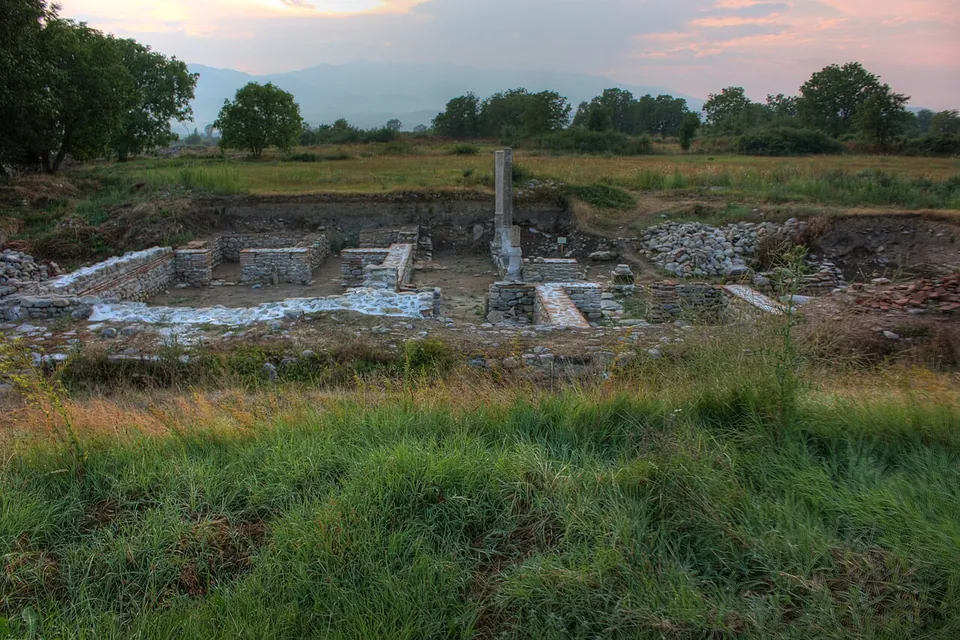 Excavated foundations of a building with low walls, white stone paving and a single standing column, in a grassy field at dusk, with trees and mountains behind.