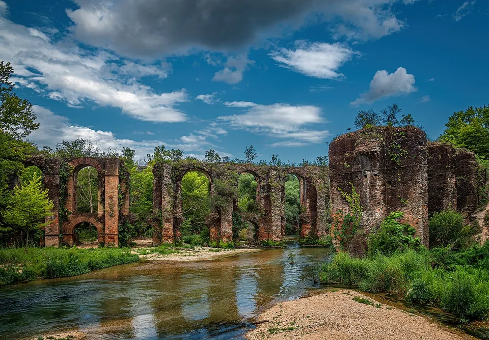 Two long lines of brick arches on tall piers crossing a shallow river among green trees, reflected in the water, under a cloudy blue sky.