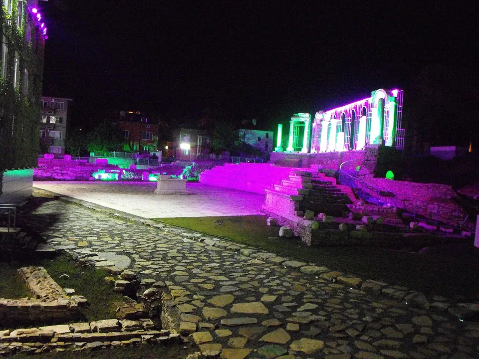 The forum at night, a paved square and a stone-paved street in the foreground, with the stone seating and a row of columns behind it lit in purple and green.