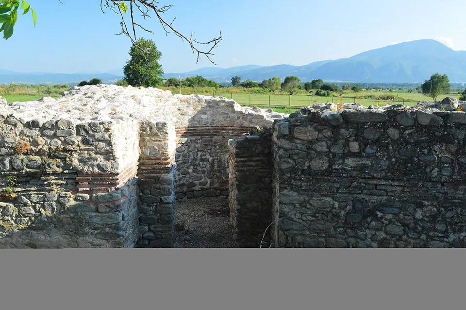 Ruined walls of rough stone with bands of brick enclose small rooms and a narrow passage, with fields, trees and mountains behind under a blue sky.