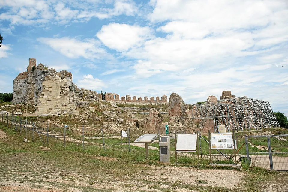 The ruined brick and stone stage building of a large theatre on a grassy slope, part of it held up by a steel lattice frame, with information boards on a fenced path in the foreground.
