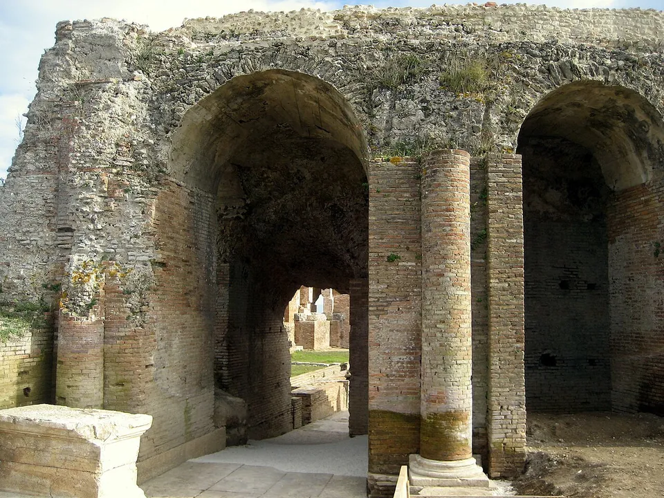 Tall arches of brick and rubble with a brick column between them, opening onto a passage paved in stone, with a stone block in the foreground.