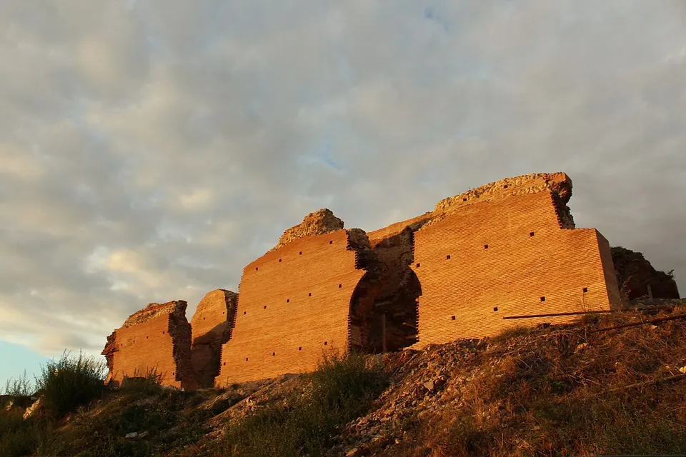 A long, high wall of red brick with a broken arch, glowing in low evening sun on top of a scrubby slope under a cloudy sky.