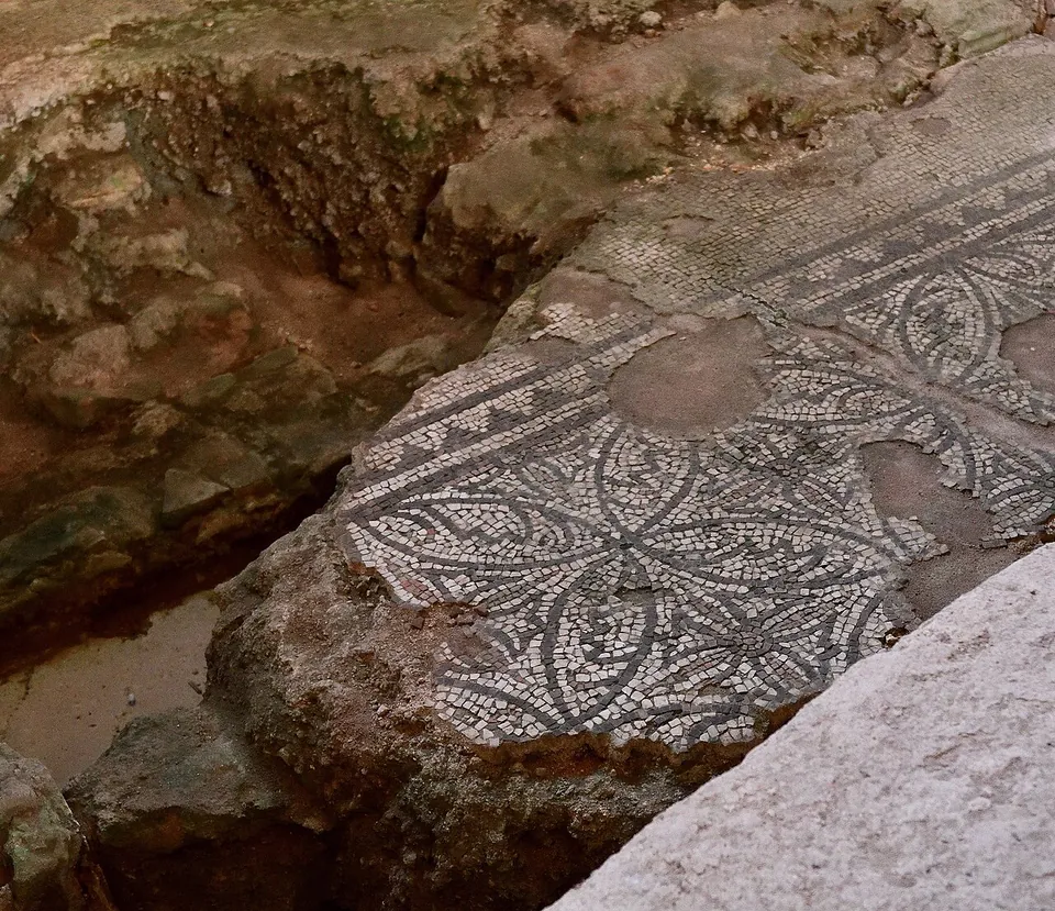 A section of mosaic floor with a geometric pattern of interlocking circles and leaf shapes in dark and light tesserae, broken at the edges, lying below the present floor level.