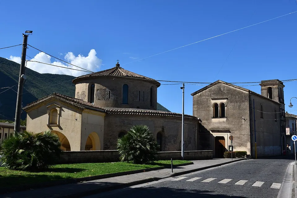A round building of pale stone with a low drum and a tiled conical roof, a lower ring and a gabled porch in front, set behind a lawn and palms on a quiet street, with a plain gabled building to the right and wooded mountains behind.