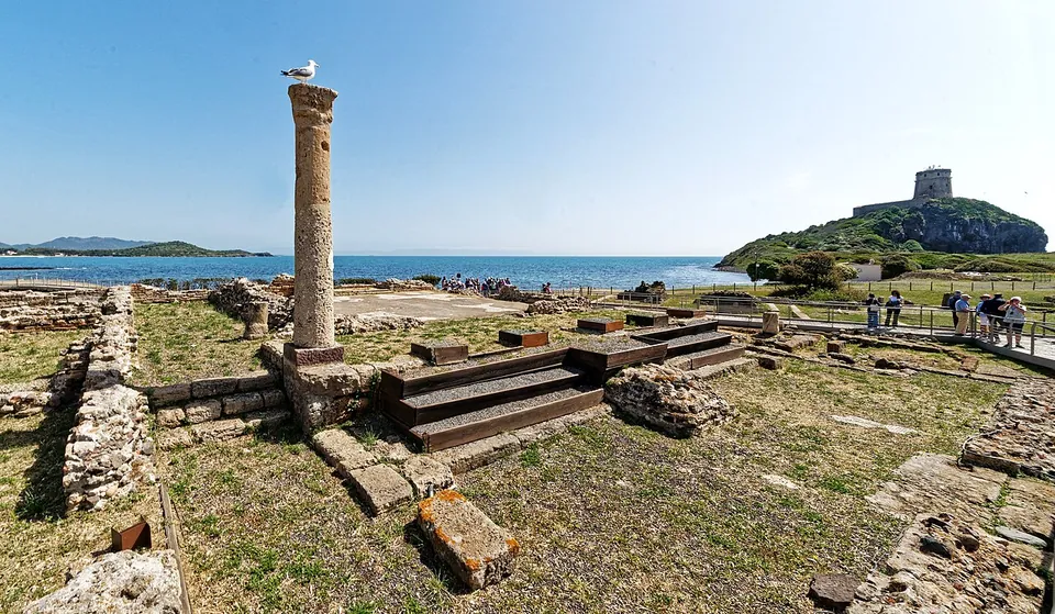 A single tall stone column on a stepped base stands among low excavated walls and grass, with visitors on a boardwalk, the sea beyond and a watchtower on a rocky headland to the right.