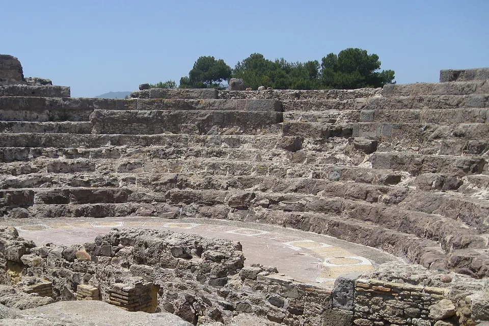 The curved stone seating of a small theatre rising in tiers above a flat semicircular orchestra, with the low walls of the stage building in the foreground and pines along the top.