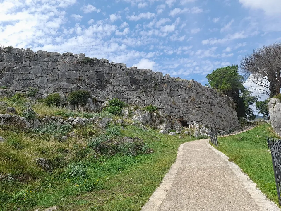 Low stone walls of excavated rooms laid out on a grassy slope, several of them under white protective covers, with a terrace wall behind and a wide plain stretching to the horizon.