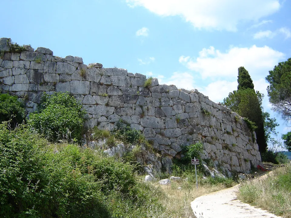 A paved path climbing beside a long, high wall of large irregular stone blocks, with bushes at its foot and trees at the far end under a blue sky.