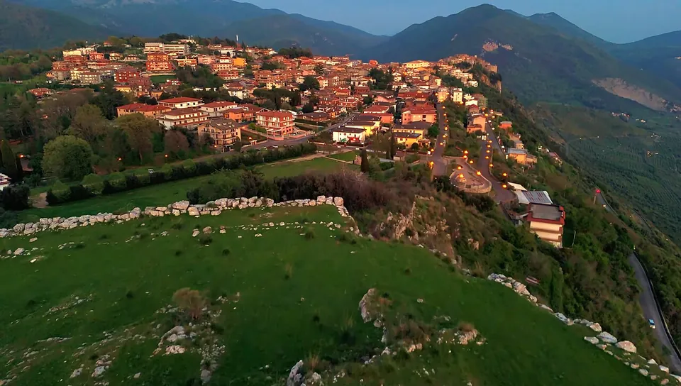 An aerial view at dusk over a green hilltop crossed by a line of pale stone blocks, with the streets and houses of a hill town spread just beyond and mountains behind.