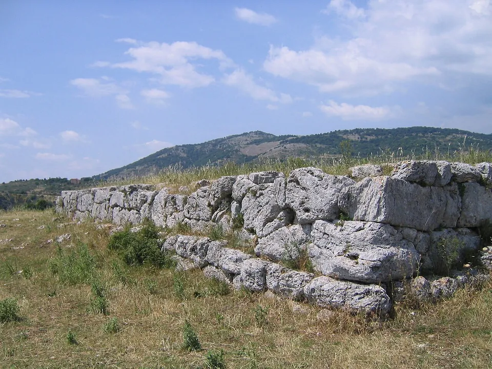 A low platform of large, roughly shaped limestone blocks in two courses, standing in dry grass with wooded hills behind.