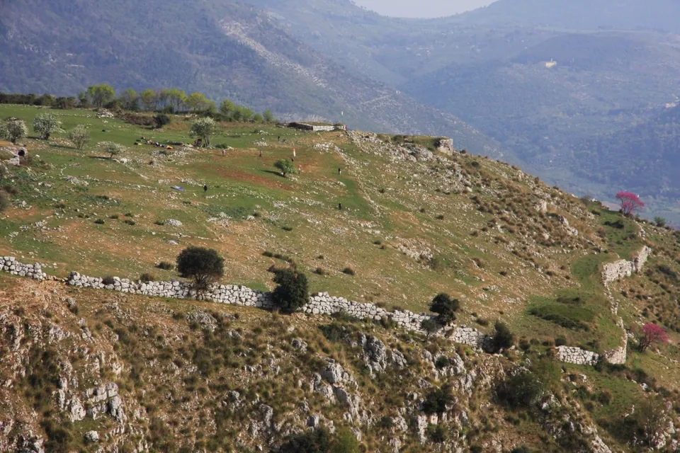 A grassy plateau falling away to a rocky slope, with a long low line of pale stone wall running across the hillside below it and further stretches of wall at the right, people walking on the plateau and mountain valleys behind.