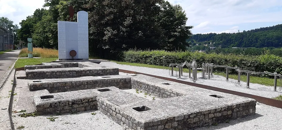 Low walls of stone laid out on gravel in the plan of a gateway, with a tall white panel, a line of metal railings and trees behind.