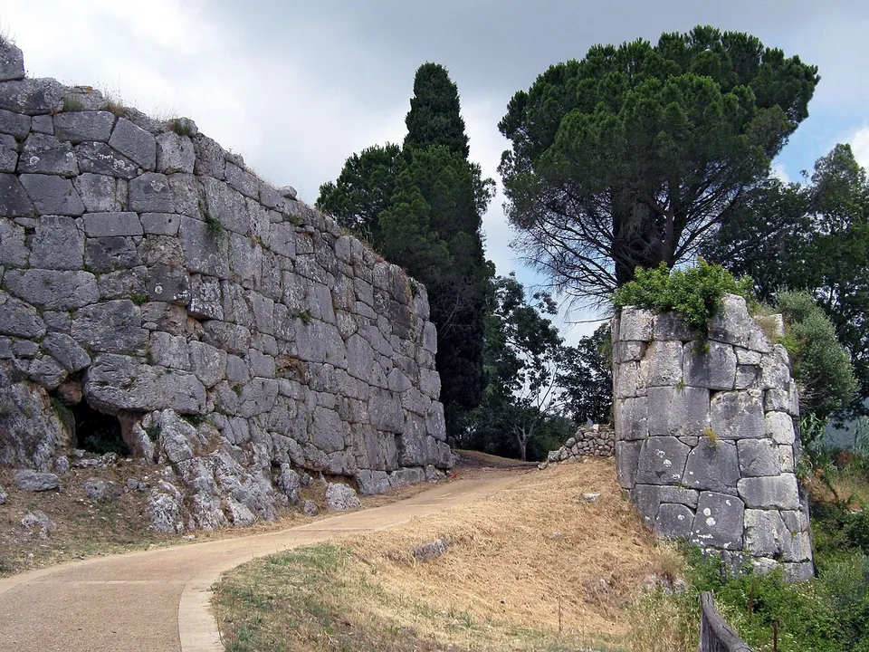 A paved path curving between a towering wall of huge, irregular grey limestone blocks fitted without mortar on the left and a rounded tower of the same masonry on the right, with pines and cypresses beyond.