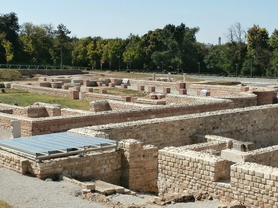 Restored low walls of pale squared stone with brick courses, forming rooms and a large rectangular hall across an open site, with a modern glass panel over one part and a line of trees behind.