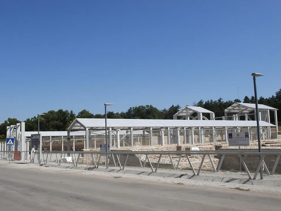 A row of modern white canopies on steel posts over excavated walls beside a road, with a pedestrian crossing sign, street lamps and trees under a clear blue sky.
