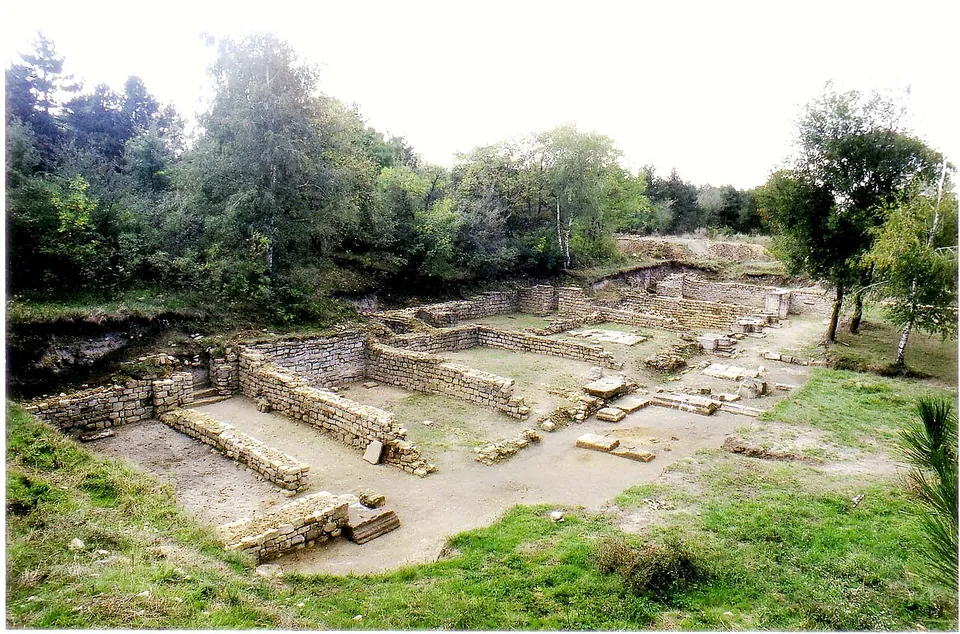 Excavated stone walls of a large building with several rooms on a terraced, grassy site, surrounded by trees.