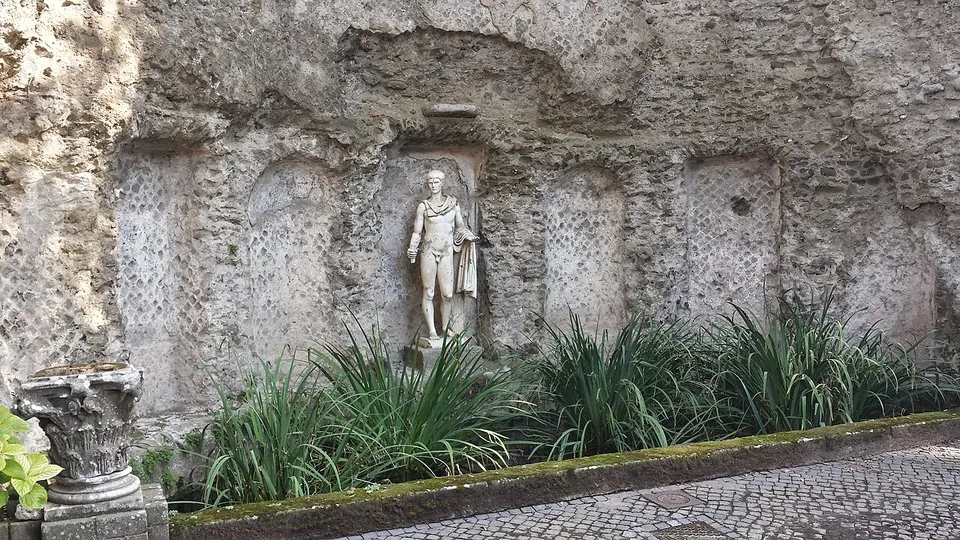 A rough rubble wall with a row of shallow arched and rectangular niches, some faced with a net-like pattern of small square stones. A marble statue stands in the central niche above a bed of spiky plants, and a carved capital rests on the ground to the left.