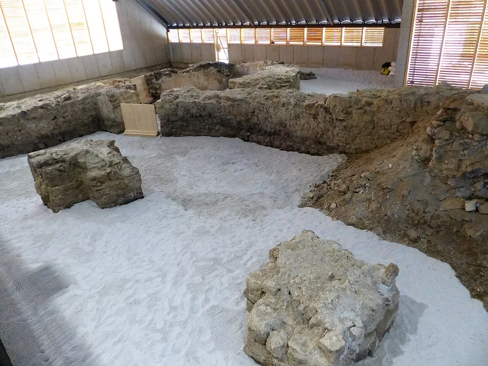 Walls of rough stone and two square blocks standing in a gravel-covered courtyard inside a modern hall, with a band of windows along the far wall.