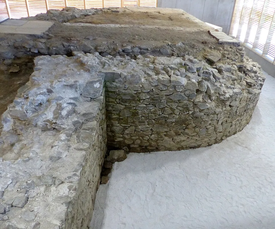 The curved wall of a round tower in rough stone masonry, a few metres high, joined to a straight wall, standing on pale gravel inside a hall with slatted wooden walls.