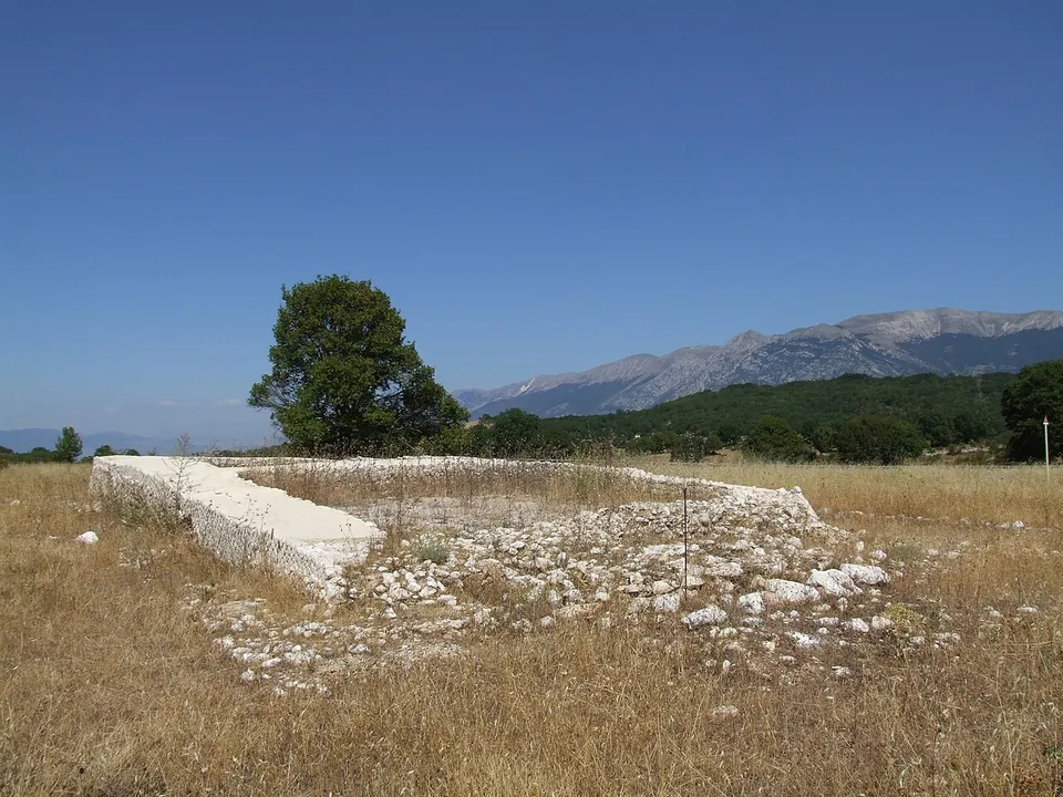 A rectangle of low, pale stone walls in dry summer grass, one end raised slightly higher than the rest, with a tree behind and a long mountain ridge on the horizon.