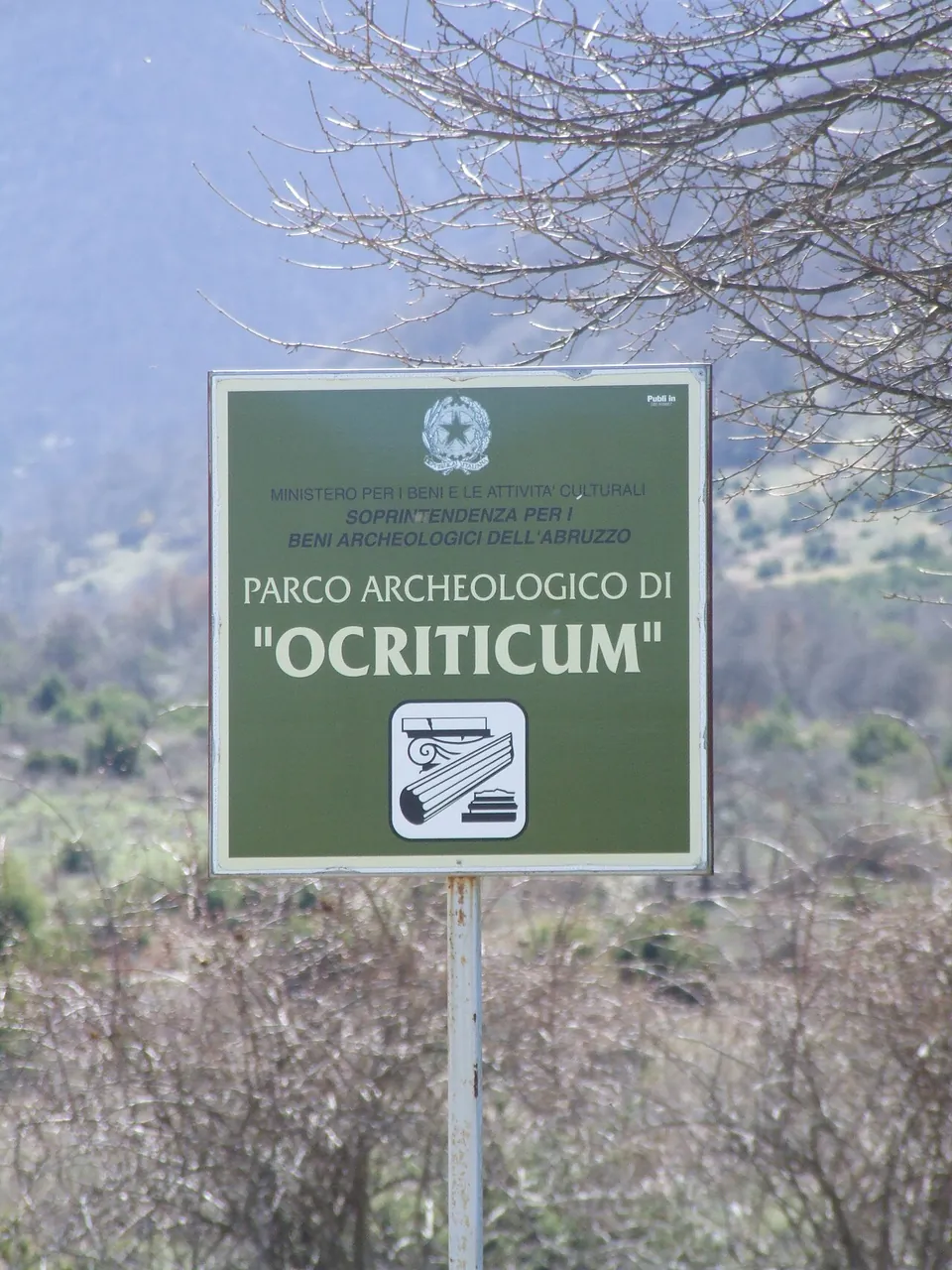 A green ministry sign on a post among bare shrubs reading "Parco archeologico di Ocriticum", under the heading of the archaeological superintendency of Abruzzo.