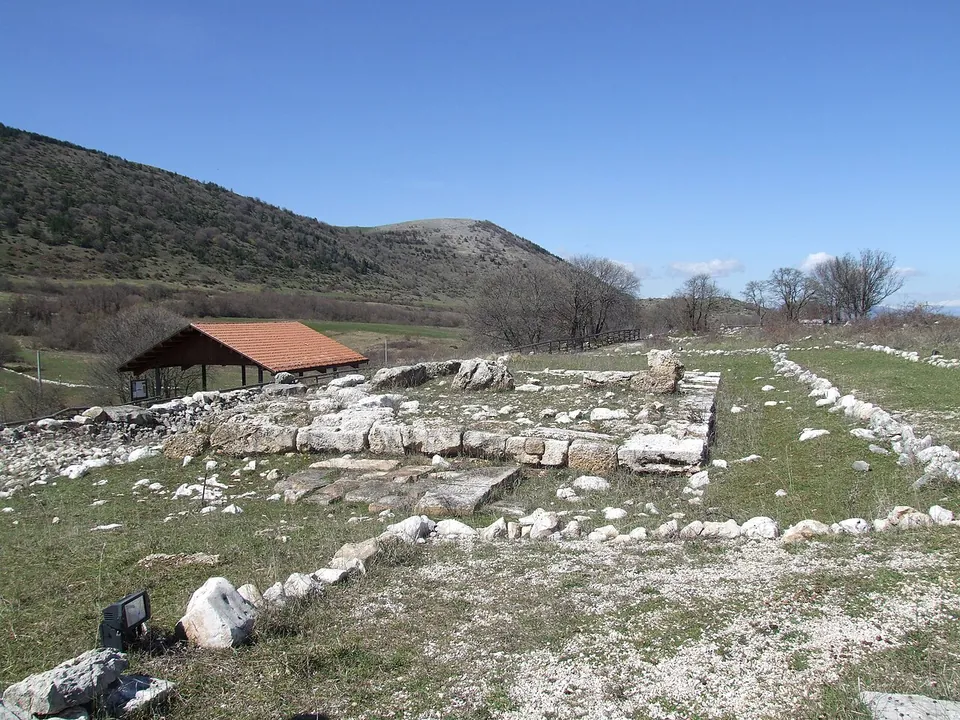 The stone foundation of a small rectangular building on a grassy terrace, with low outline walls running away from it and a timber shelter with a red tiled roof beyond, under a hillside.