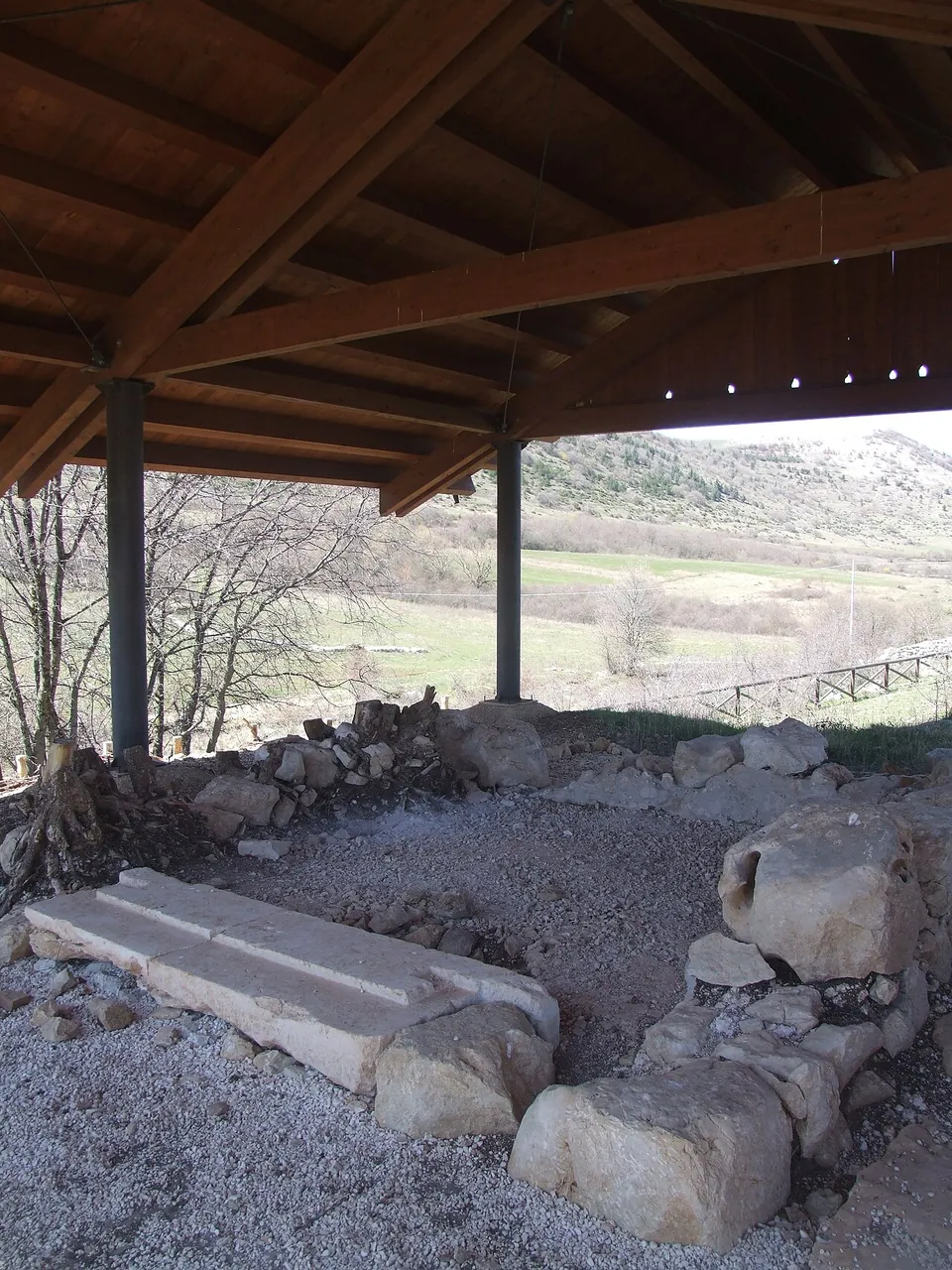Under a timber roof on posts, a small square of rough stone walls on a gravel floor, with long dressed blocks lying at its front and open country beyond.
