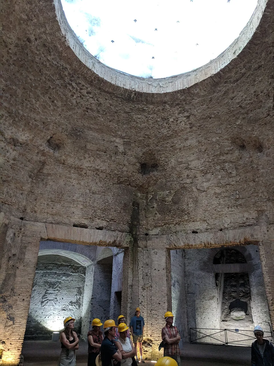 Looking up the rough concrete wall of a dome to a large round opening full of white sky, with visitors in hard hats grouped below and lit doorways into the side rooms.