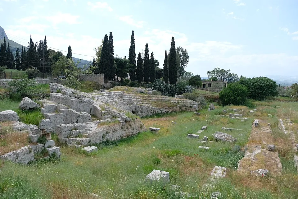 The curved stone seating of a small theatre set into a grassy slope, with cypresses above and scattered blocks in the grass in front.