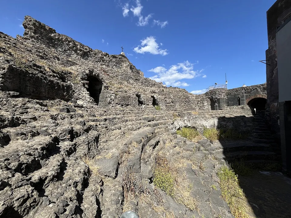 Rough rows of black lava seating curving round inside the Odeon, with dark openings in the wall at the top and an arched passage at the far end.