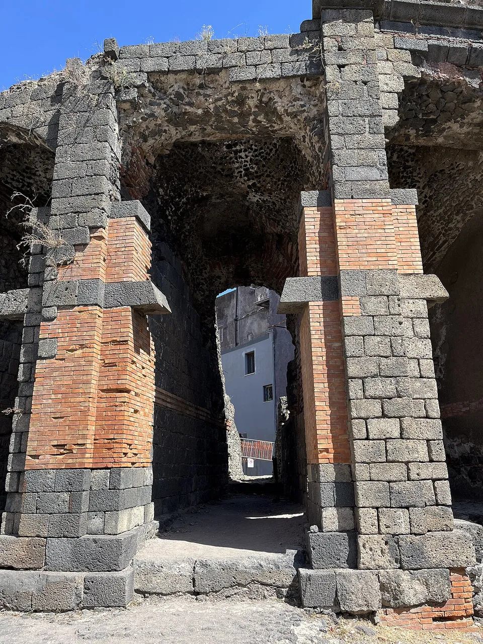 A narrow vaulted passage between two piers of grey lava blocks banded with red brick, with a white building visible through the far end.