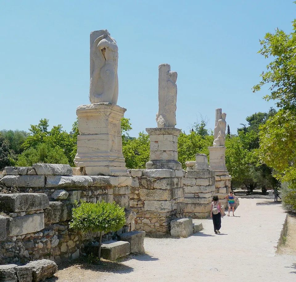 Three tall marble statues on high square pedestals along a wall of stone blocks beside a sunlit path, the nearest a figure with a fish's tail, the others worn, with trees behind and two visitors walking past.