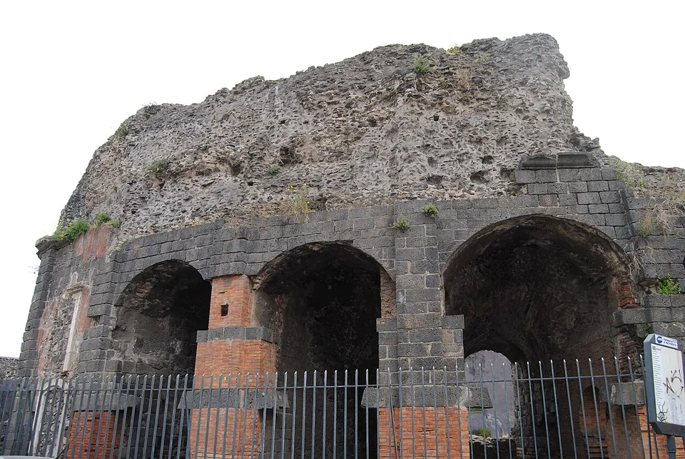 The curved outer wall of the Odeon, built of black lava blocks, opening in a row of tall arches whose piers are faced with red brick, with a rough mass of masonry rising above and a railing in front.