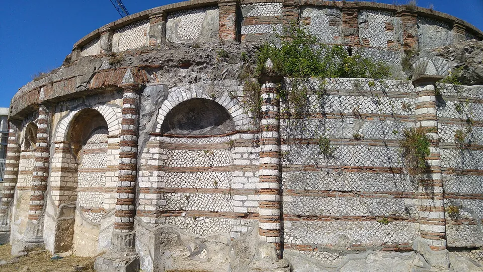 A stretch of the outer wall with two round-headed niches and pilasters banded in brick, the wall between them faced with small square stones laid in a diagonal net pattern, under a crumbling upper edge.