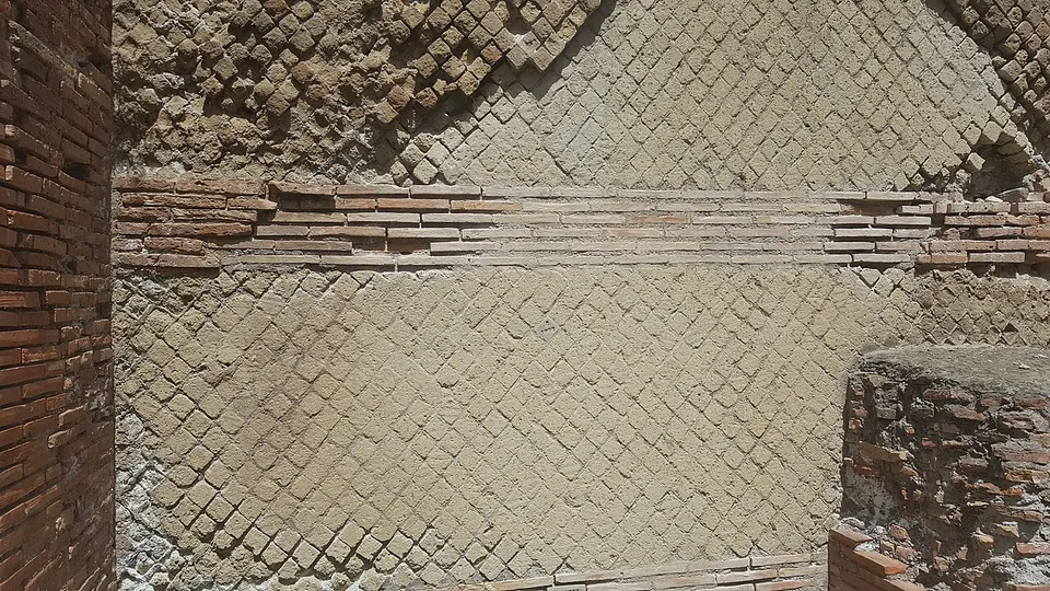 A wall faced with small square stones set diagonally in a net pattern, crossed by a band of thin red bricks, between two brick piers.