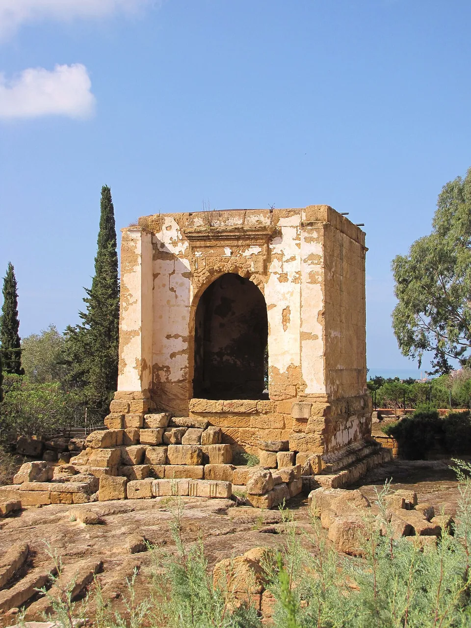 A small square building of pale stone with a large round-arched opening in its front wall, standing on a stepped base among cypress trees.