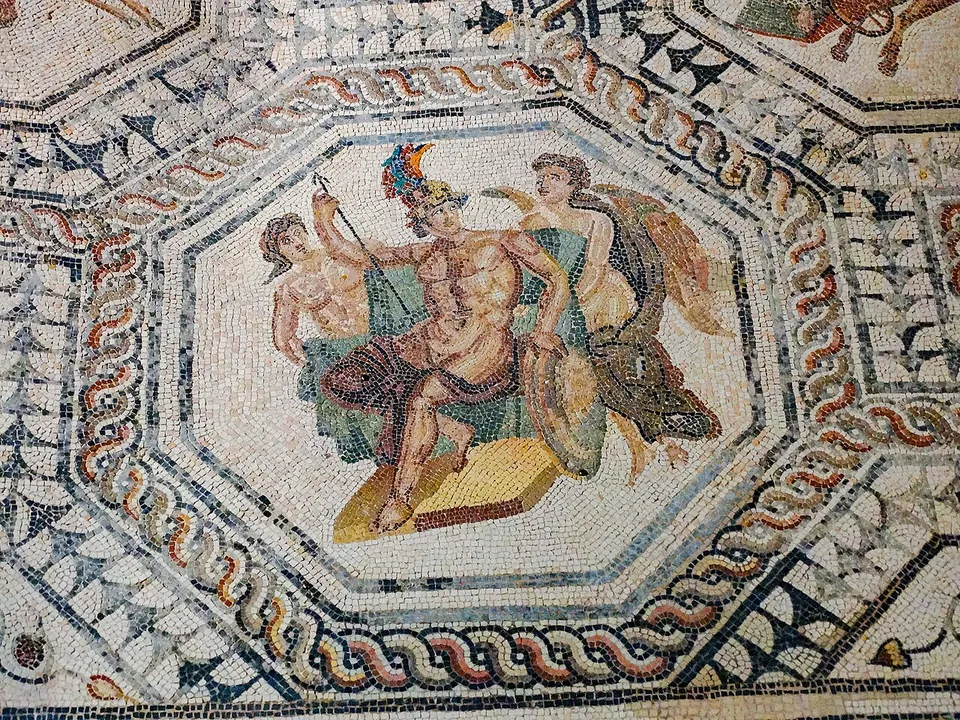 An octagonal mosaic panel framed by braided borders, showing a man in a crested helmet holding a staff and a shield, with two other figures beside him, set in a floor of geometric patterns.