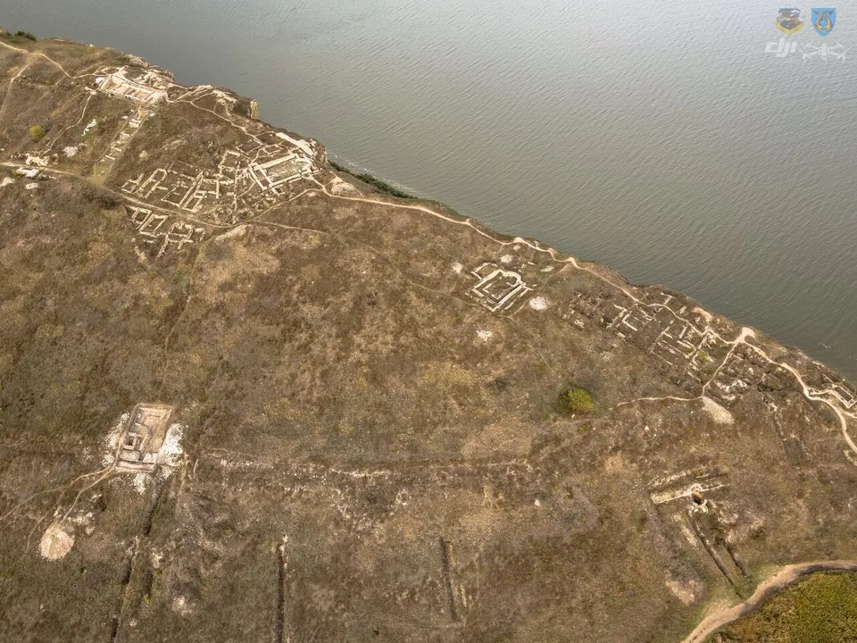 An aerial view along a low cliff above a wide lake, with the stone outlines of excavated buildings and walls spread over the dry grassland of the headland.