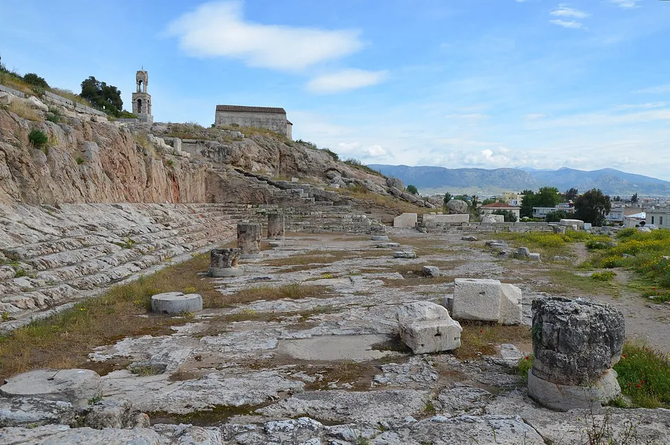 A wide flat expanse of rock-cut floor and column bases at the foot of a rocky hill, with rows of seats cut into the rock along one side and a church and bell tower on the hill above.