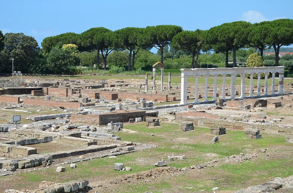 A broad field of excavated stone foundations and low walls in mown grass, with a row of white columns carrying an architrave on the right and a line of umbrella pines behind.
