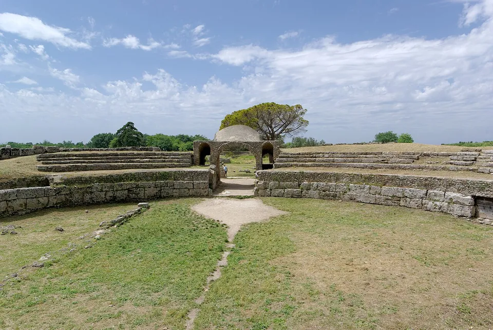 A grassy oval arena ringed by a low stone wall, with a few rows of stone seating above it and a vaulted entrance passage at the far end.