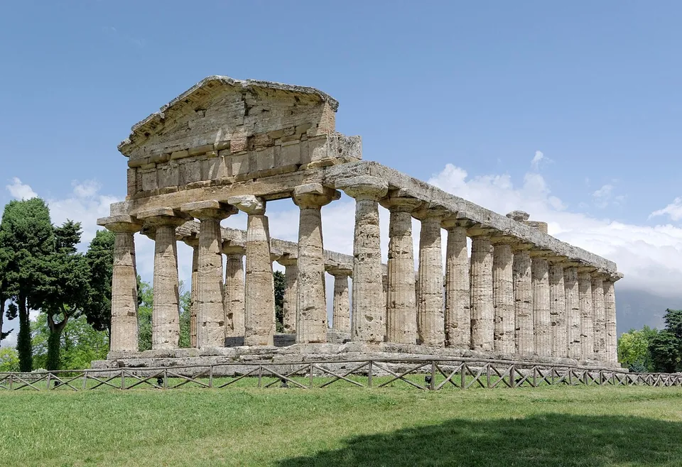 A smaller Greek Doric temple with a surviving triangular gable at its front end and a line of columns along its side, among trees under a blue sky.