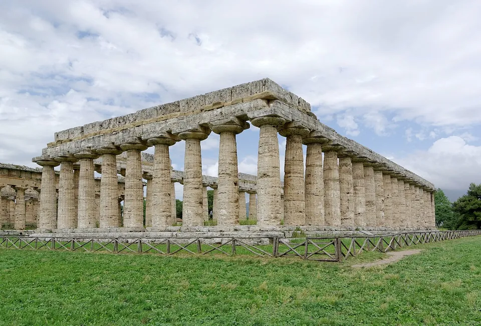 A long Greek temple of weathered stone columns with broad cushion-shaped capitals, carrying only a single course of stone beams and no roof or gable, seen across a lawn.