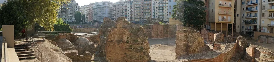 A very wide panorama of an excavated site of brick walls and foundations below a modern square, with steps and railings at the edge and apartment buildings all round.