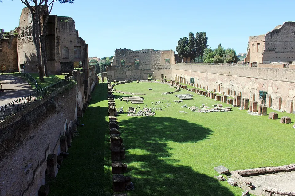 A long sunken lawn seen from above at one end, enclosed by high brick walls, with rows of pier bases along its sides, scattered marble fragments on the grass and ruined brick buildings beyond.