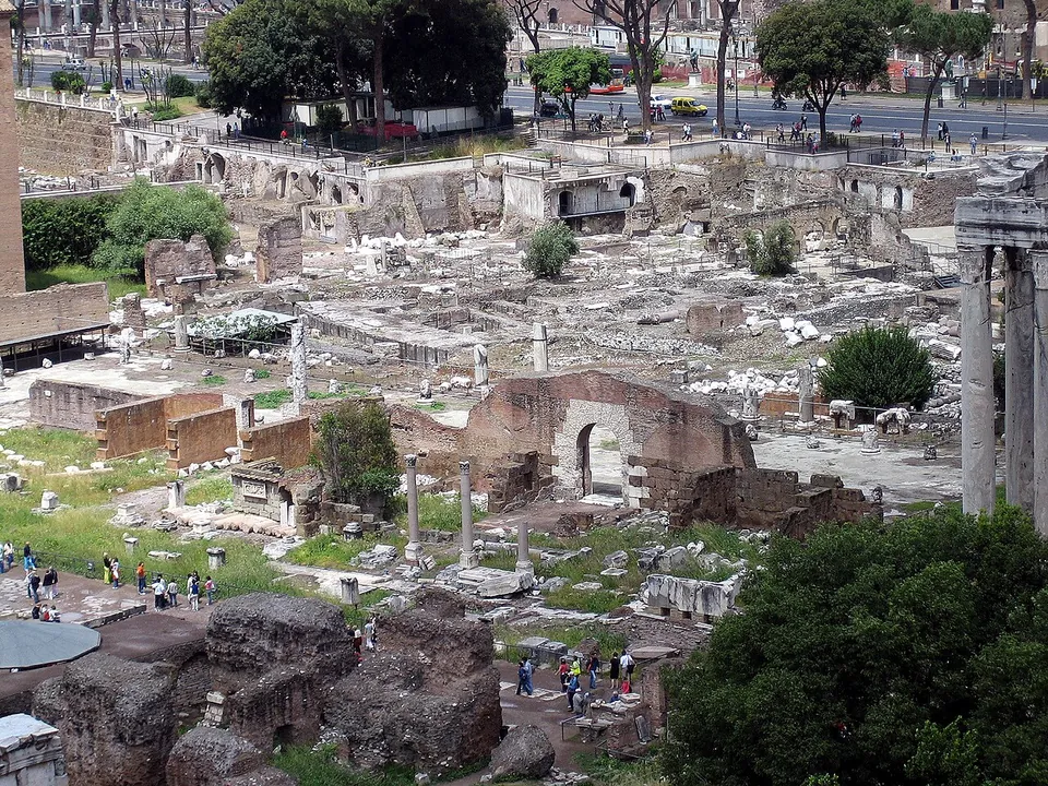 Looking down from a height over a field of ruins, low walls, column stumps and marble fragments with paths and visitors among them, the columns of a temple at the right edge and a modern road with parked cars at the top.