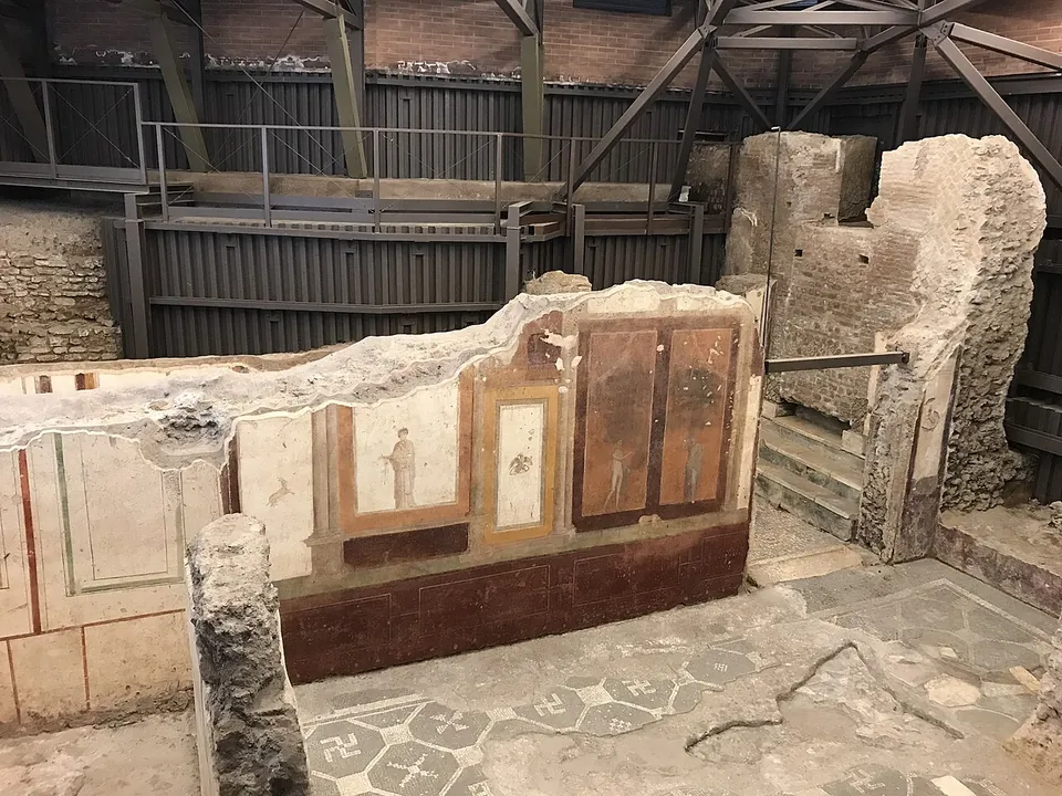 Low walls under a modern steel roof, one of them still painted with red and white panels holding small figures, beside a floor of grey and white mosaic in a pattern of hexagons.