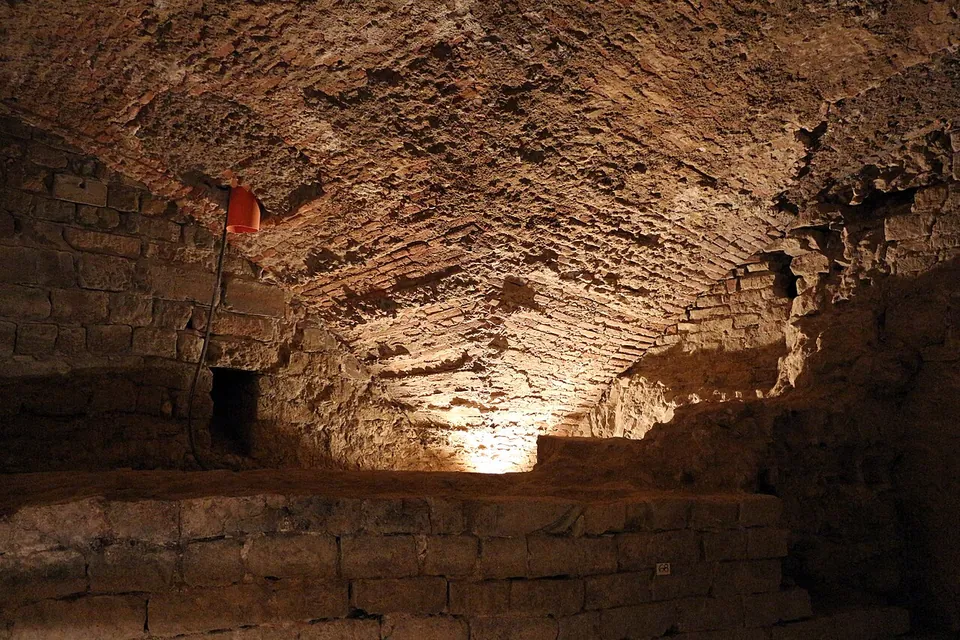 A low underground chamber under a rough brick barrel vault, lit from below, with a ledge of squared stone blocks running along the floor in front of it.