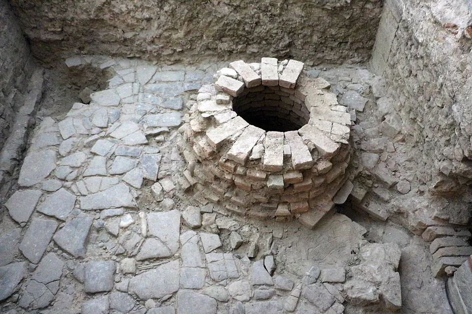 Looking down into an excavated pit floored with irregular stone paving, with a round well-head of stacked bricks in the middle and cut earth walls around it.